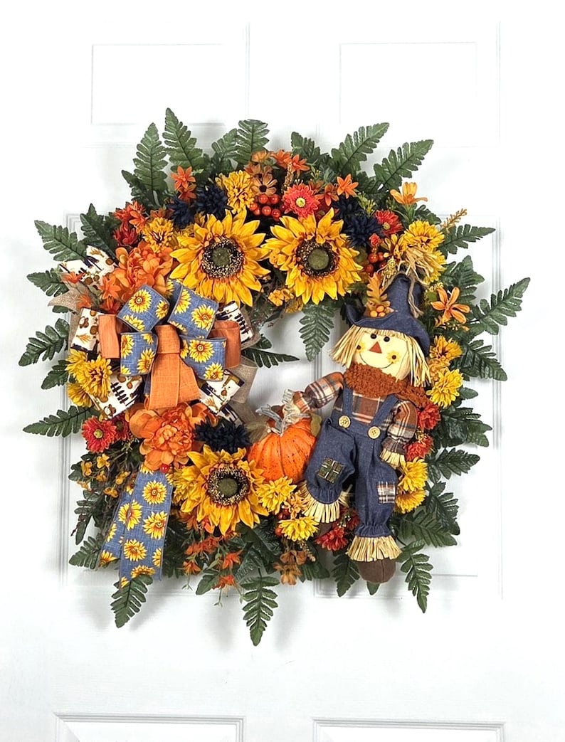 Puede incluir: Una corona decorativa de oto&ntilde;o con girasoles amarillos, flores naranjas, hojas de helecho verdes y una peque&ntilde;a calabaza naranja. Incluye una figura de espantap&aacute;jaros de tela con sombrero y peto azul, decorada con un lazo azul estampado.