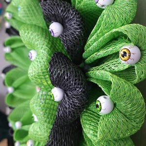 May include: A green and black Halloween wreath with multiple googly eyes attached. The wreath is made of mesh and is decorated with black and green bows.