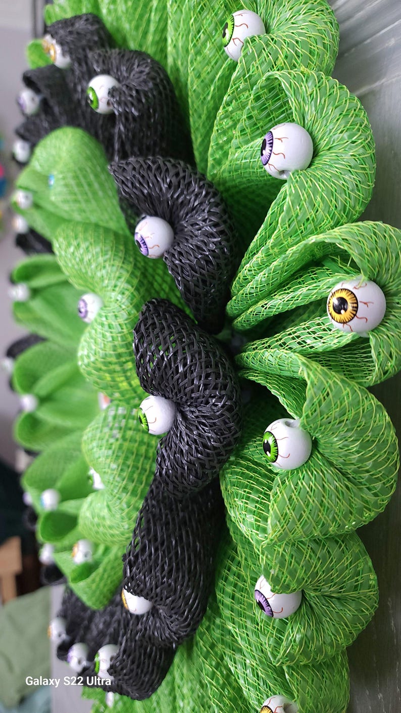 May include: A green and black Halloween wreath with multiple googly eyes attached. The wreath is made of mesh and is decorated with black and green bows.
