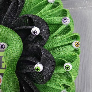 May include: A green and black mesh wreath decorated with multiple colourful googly eyes. The wreath is a Halloween decoration.