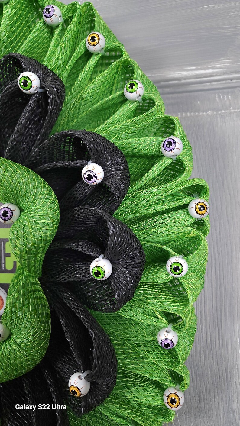 May include: A green and black mesh wreath decorated with multiple colourful googly eyes. The wreath is a Halloween decoration.