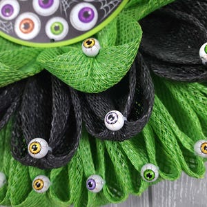 May include: A green and black mesh wreath decorated with googly eyes. The wreath has a sign that says "Welcome" in white lettering.