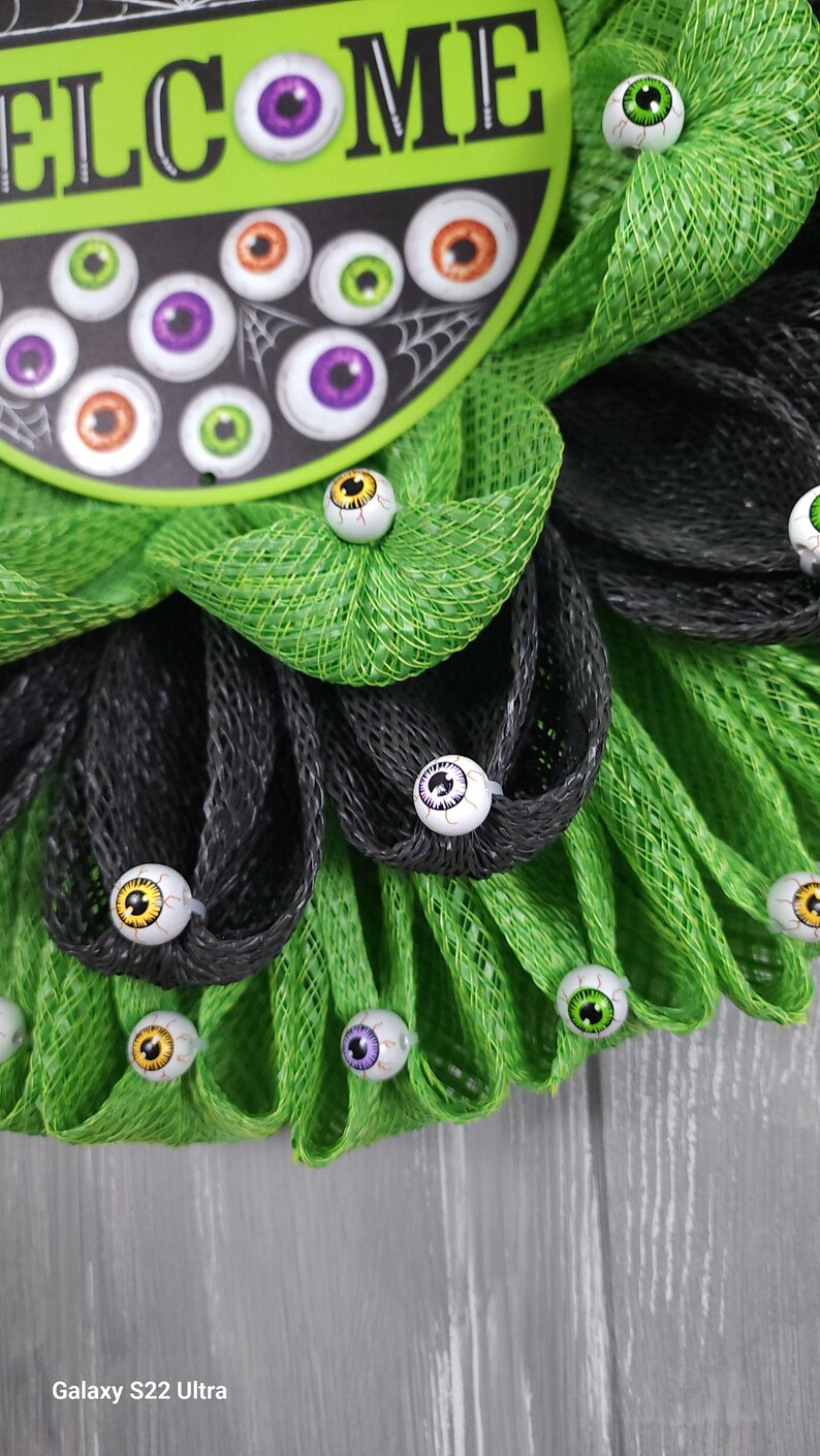 May include: A green and black mesh wreath decorated with googly eyes. The wreath has a sign that says "Welcome" in white lettering.