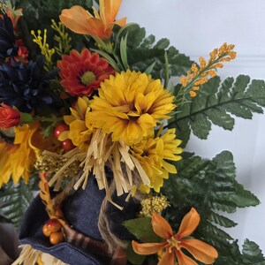 Puede incluir: Una corona decorativa con una figura de espantap&aacute;jaros que lleva un sombrero azul oscuro, rodeada de follaje oto&ntilde;al artificial que incluye crisantemos amarillos, lirios naranjas, flores azul oscuro, hojas de helecho verdes y bayas rojas.