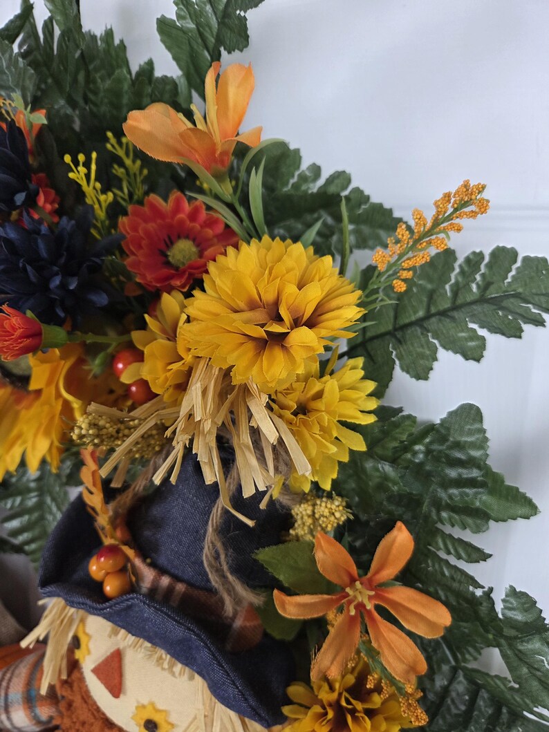 Puede incluir: Una corona decorativa con una figura de espantap&aacute;jaros que lleva un sombrero azul oscuro, rodeada de follaje oto&ntilde;al artificial que incluye crisantemos amarillos, lirios naranjas, flores azul oscuro, hojas de helecho verdes y bayas rojas.