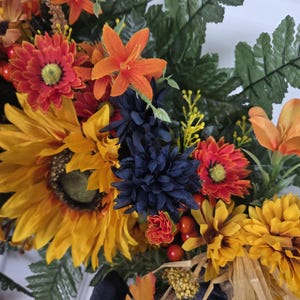 Puede incluir: Una corona decorativa que presenta una mezcla de girasoles artificiales, margaritas naranjas, crisantemos azul oscuro y hojas de helecho verdes. El arreglo incluye peque&ntilde;as bayas naranjas, flores amarillas y acentos de rafia natural.