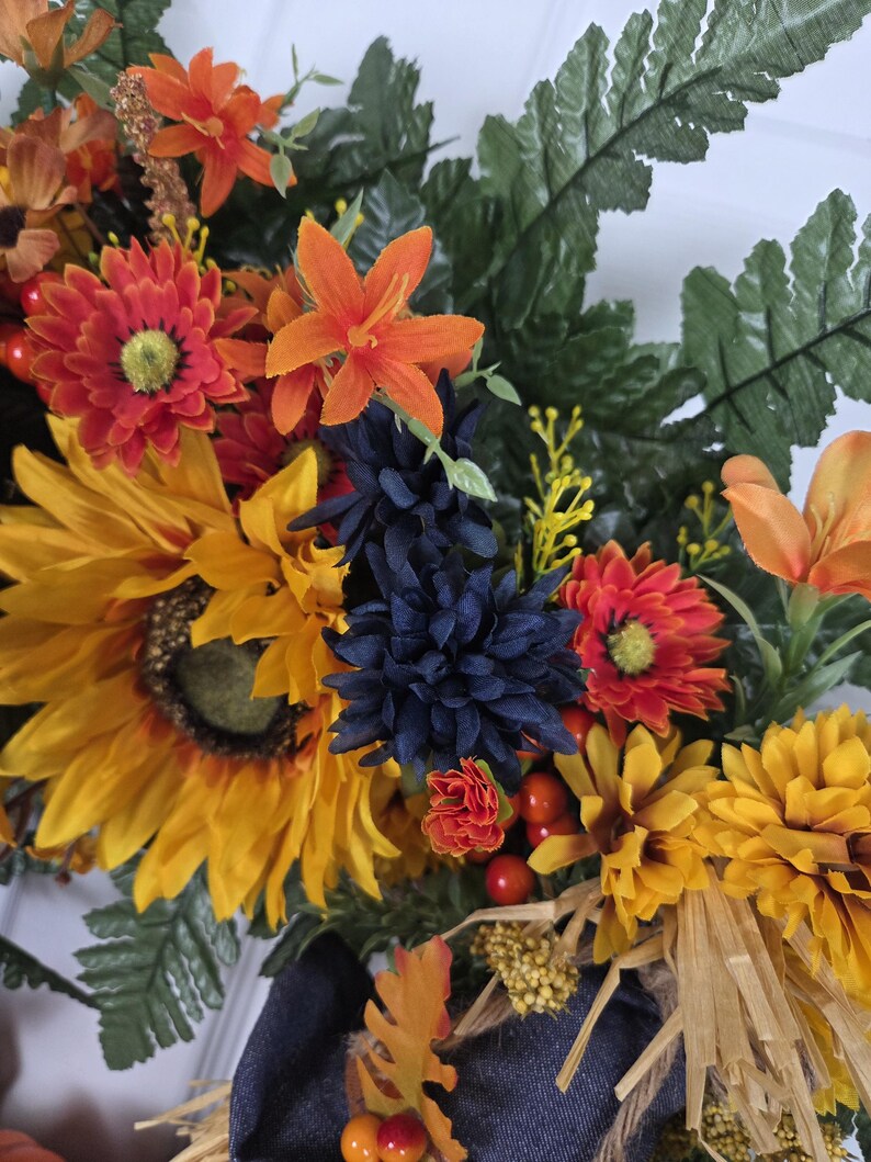 Puede incluir: Una corona decorativa que presenta una mezcla de girasoles artificiales, margaritas naranjas, crisantemos azul oscuro y hojas de helecho verdes. El arreglo incluye peque&ntilde;as bayas naranjas, flores amarillas y acentos de rafia natural.