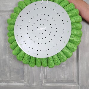 May include: A green wreath with a metal base and a white string for hanging. The wreath is made of mesh and has a circular shape. The metal base has a pattern of holes. The wreath is hanging on a grey wooden door. Galaxy S22 Ultra.