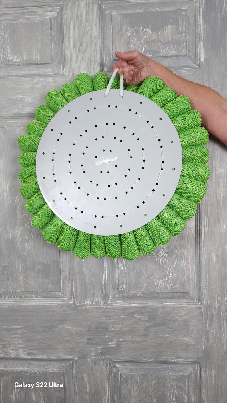 May include: A green wreath with a metal base and a white string for hanging. The wreath is made of mesh and has a circular shape. The metal base has a pattern of holes. The wreath is hanging on a grey wooden door. Galaxy S22 Ultra.