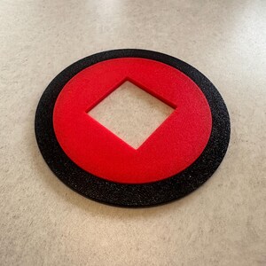 Peut inclure: Un dessous de verre circulaire avec un anneau extérieur noir, un anneau intérieur rouge et une découpe en forme de losange rouge au centre. Le dessous de verre est fait d'un matériau texturé.