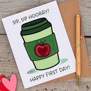 May include: A white greeting card featuring a green coffee cup illustration with a red apple graphic, the text "SIP, SIP HOORAY!" and "HAPPY FIRST DAY!", accompanied by a wooden pen and two small paper hearts on a rustic wooden surface.
