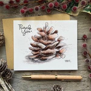 May include: A white card with the words "Thank You" written in black ink. A brown pine cone is drawn in the center of the card. The card is sitting on a wooden surface with a gold ribbon and red berries.
