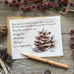 May include: A greeting card with a watercolor illustration of a pine cone and the text "Pine cones are little treasures that drop from the trees...all different, all beautiful, all perfect in our imperfection." A wooden pen rests on the surface.