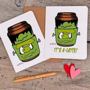 May include: Two white greeting cards featuring a cartoon illustration of a green coffee cup designed to look like Frankenstein's monster, complete with bolts and stitches. The text on one card reads IT'S A-LATTE! with a wooden pen and two paper hearts.
