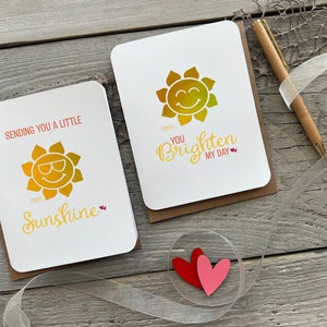 May include: Two greeting cards with a sun design. The first card says "Sending you a little sunshine." The second card says "You brighten my day."