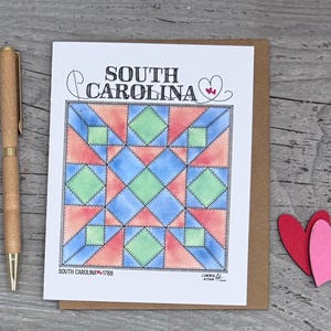 May include: A greeting card featuring a watercolor quilt design in shades of blue, orange, and green, with the words "SOUTH CAROLINA" at the top. A wooden pen and two heart-shaped cutouts are next to the card.