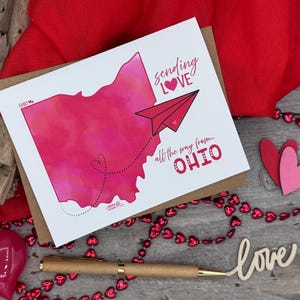 May include: A white card with a pink watercolor illustration of the state of Ohio. A red paper airplane flies out of the state with the text "sending love" and a heart. The text "all the way from... OHIO" is below the airplane.