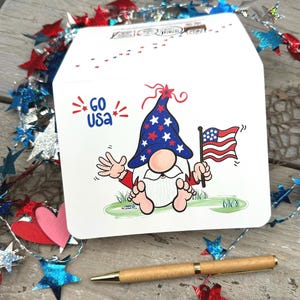 Showcasing the front of the card and glimpses of its back, this charming patriotic gnome proudly waves an American flag and his hand amidst red, white, and blue colors.