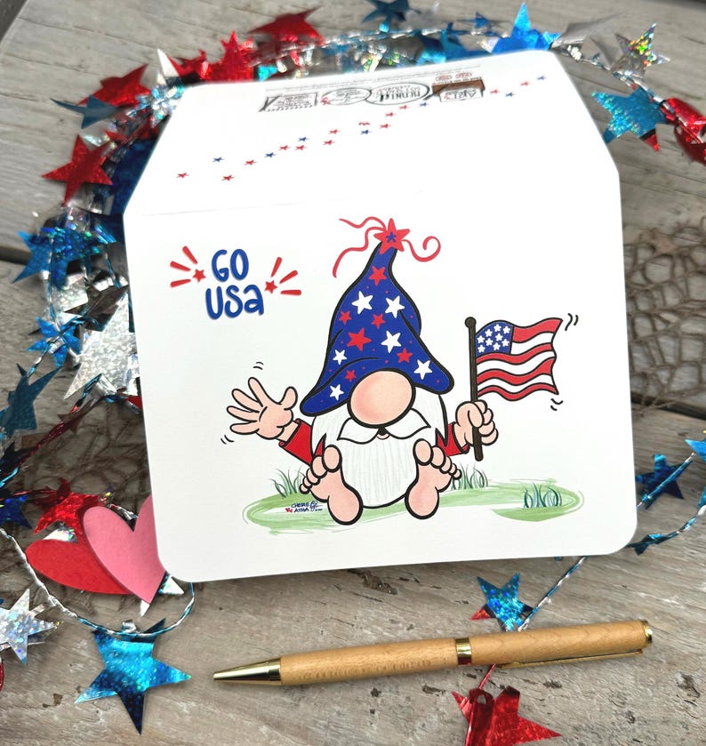Showcasing the front of the card and glimpses of its back, this charming patriotic gnome proudly waves an American flag and his hand amidst red, white, and blue colors.
