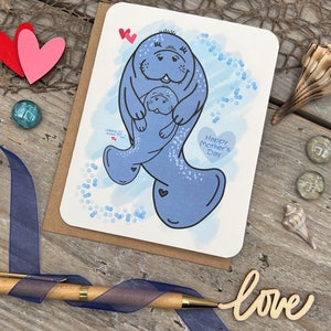 Printed on 80 lb white card stock this Mothers Day card has two original illustrated manatees that represents a momma and child. The larger manatee is holding the smaller manatee in its flippers. It has a blue digital watercolor background with bubbles to show them swimming in the ocean. The card has the text Happy Mothers Day along with my signature hearts.