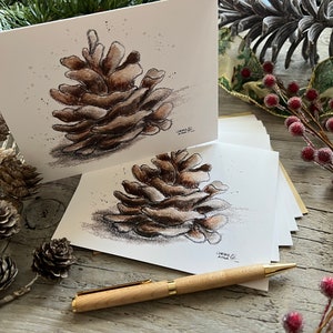 May include: Two white cards with a hand-drawn illustration of a brown pine cone on each. The cards are on a wooden surface with a pen and pine cones.