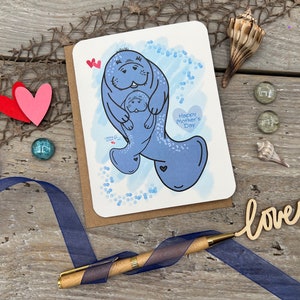 A note card for Mothers Day with a blue manatee and a baby manatee on front. In the background is a blue digital watercolor with blue bubbles. The mom manatee is holding the younger manatee. The card has the text Happy Mother's Day in a heart shape.
