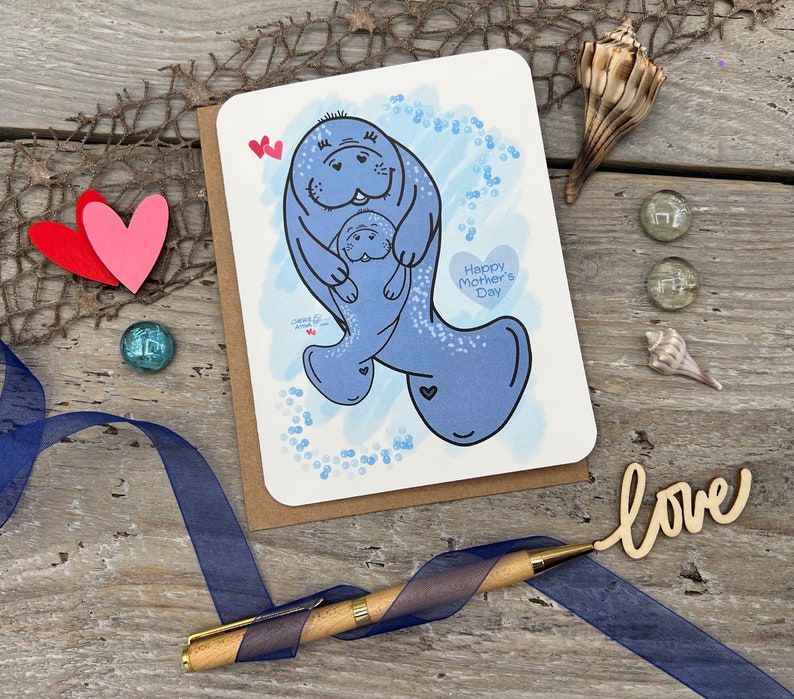 A note card for Mothers Day with a blue manatee and a baby manatee on front. In the background is a blue digital watercolor with blue bubbles. The mom manatee is holding the younger manatee. The card has the text Happy Mother's Day in a heart shape.