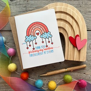 May include: A white card with a rainbow, clouds, and hearts. The text on the card reads "Thank you for being my rainbow throughout my storm."