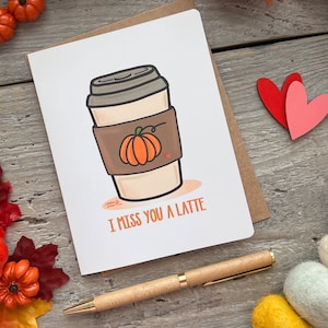 May include: Greeting card with a coffee cup illustration featuring a pumpkin design. The card reads "I MISS YOU A LATTE" in orange text. A wooden pen and decorative pumpkins are also visible.
