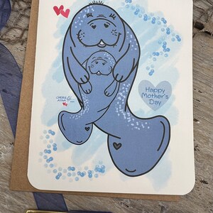 This card is an original hand drawn image of a momma and young manatee. The greeting card has a blue watercolor background and blue bubbles. The bigger manatee represents the mother and the smaller one represents a baby or younger manatee. The text on the card reads Happy Mothers Day.