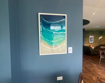 May include: Framed abstract artwork of a beach scene with ocean waves. The piece uses shades of blue and turquoise, with a sandy beige shoreline. The white-framed artwork is mounted on a dark blue wall, creating a coastal theme.
