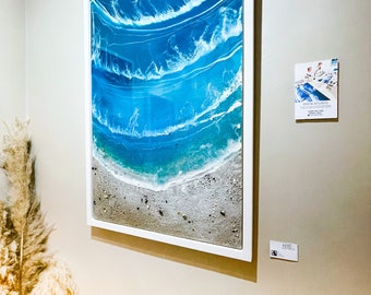 May include: A framed artwork showing an ocean scene. The art piece uses layers of blue and white resin to create the illusion of waves breaking on a sandy shore. The artwork is in a white frame and mounted on a neutral-coloured wall.