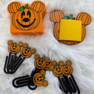 May include: Orange pumpkin-shaped desk accessories and paper clips. The pumpkin designs have Mickey Mouse ears and smiling faces. The accessories include a memo pad holder and paper clips, perfect for Halloween or fall-themed decor.