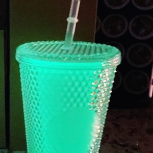 Puede incluir: Un vaso reutilizable verde que brilla en la oscuridad con un diseño texturizado y una pajita de plástico transparente.