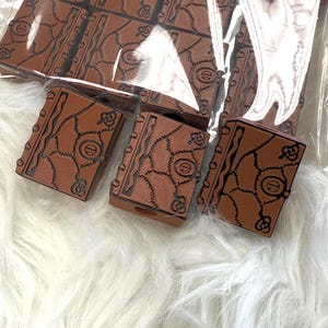 Puede incluir: Artículos decorativos rectangulares marrones con diseño de libro. Los artículos tienen un patrón detallado en relieve con detalles en negro. Los artículos están en una bolsa de plástico transparente y sobre una superficie blanca y esponjosa.