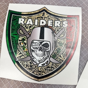 Puede incluir: Una pegatina con un diseño de escudo con la palabra "RAIDERS" en blanco. El escudo tiene un borde verde, blanco y rojo, con una calavera que lleva un casco de fútbol en el centro. Dos espadas cruzadas están a cada lado del casco.