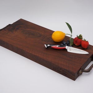 May include: A large, rectangular, dark brown wooden cutting board with two metal handles. The board has a knife, a lemon, blueberries, and strawberries on top.