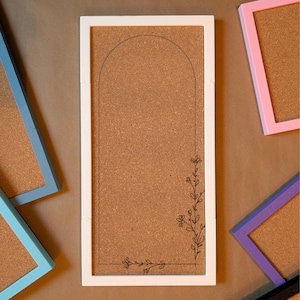 May include: A rectangular cork board with a white frame, featuring a black floral design and an arched outline. Other cork boards with blue, pink, and purple frames are visible in the background. The cork board is ideal for displaying notes and photos.