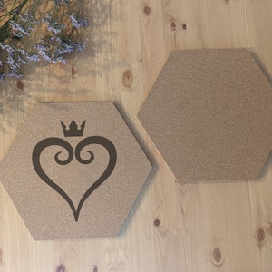 May include: Two hexagonal cork coasters. One coaster features a black heart design with a crown on top. The coasters are light brown and sit on a light wood surface. Decorative purple flowers are in the background.