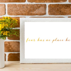 May include: White picture frame with a white mat and the text "fear has no place here" in gold lettering on a white background. The frame is sitting on a wooden surface in front of a brick wall. A vase with yellow flowers is to the left of the frame.