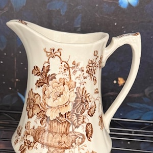 May include: Cream-colored ceramic pitcher with a brown floral design. The pitcher features a large floral arrangement in a basket, with additional floral and leaf accents. The handle is curved and the spout is wide. A decorative piece for the kitchen or dining room.