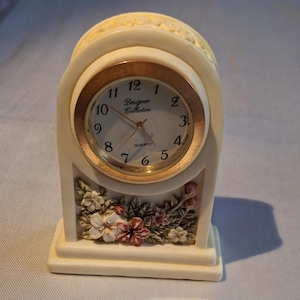 May include: An off-white mantel clock with a round face, gold-tone bezel, and black numerals. The clock face displays the text Designer Collection and Quartz. The base features a relief sculpture of multi-colored flowers and green leaves.