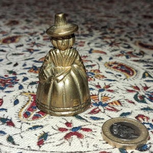 May include: A small, gold-coloured figurine of a person wearing a hat and traditional clothing. A coin is next to the figurine. The figurine is on a patterned fabric.