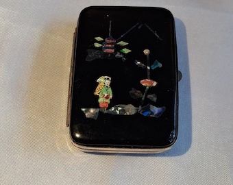 May include: A rectangular black case featuring an inlaid design of a figure in a green garment carrying a basket, a small pagoda, and a flower. The inlay is composed of iridescent, multi-colored pieces that contrast against the glossy black surface.