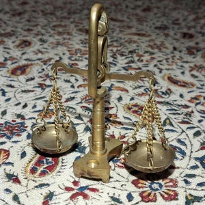 May include: A small, antique brass scale with two hanging bowls connected by chains. The scale has a central support column and a curved handle at the top. The background is a patterned fabric with floral and paisley designs.