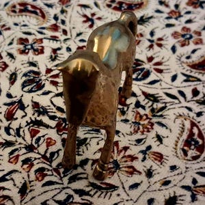 May include: A small, brass horse figurine with a polished finish. The horse is standing on four legs, with its head and neck arched. The figurine is set against a patterned fabric with red, blue, and white floral designs.