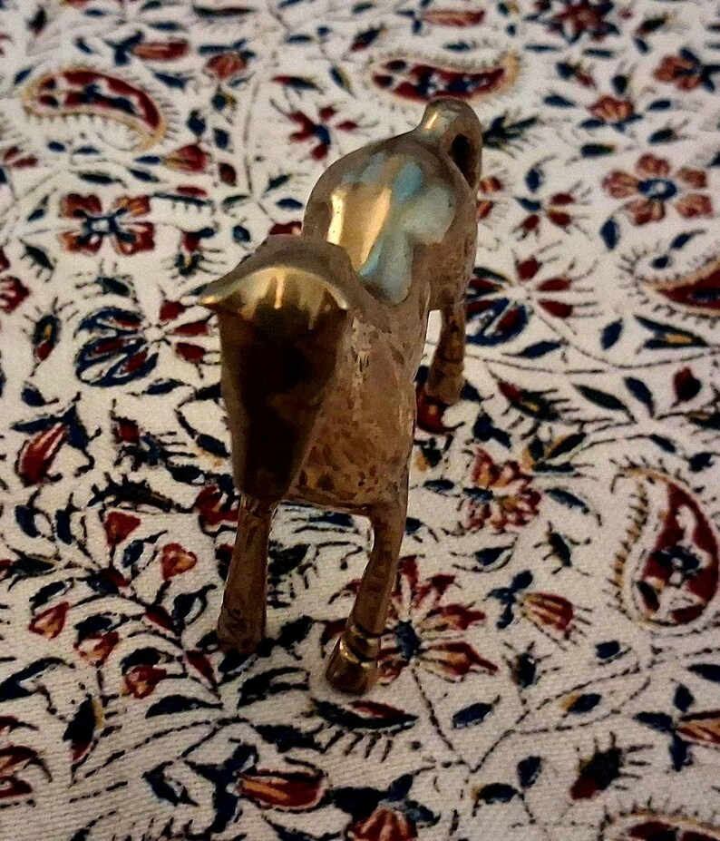 May include: A small, brass horse figurine with a polished finish. The horse is standing on four legs, with its head and neck arched. The figurine is set against a patterned fabric with red, blue, and white floral designs.