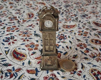 May include: A miniature silver-colored grandfather clock with a white clock face and black hands. The clock has a rectangular body with a decorative top and a window-like design on the lower portion. A coin is next to the clock.