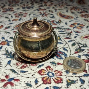 May include: A small, antique brass pot with a lid and two handles. The pot is round with a slightly tarnished finish. A coin is next to the pot for size comparison. The pot sits on a floral patterned fabric.