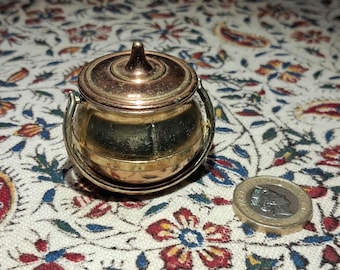 May include: A small, antique brass pot with a lid and two handles. The pot is round with a slightly tarnished finish. A coin is next to the pot for size comparison. The pot sits on a floral patterned fabric.